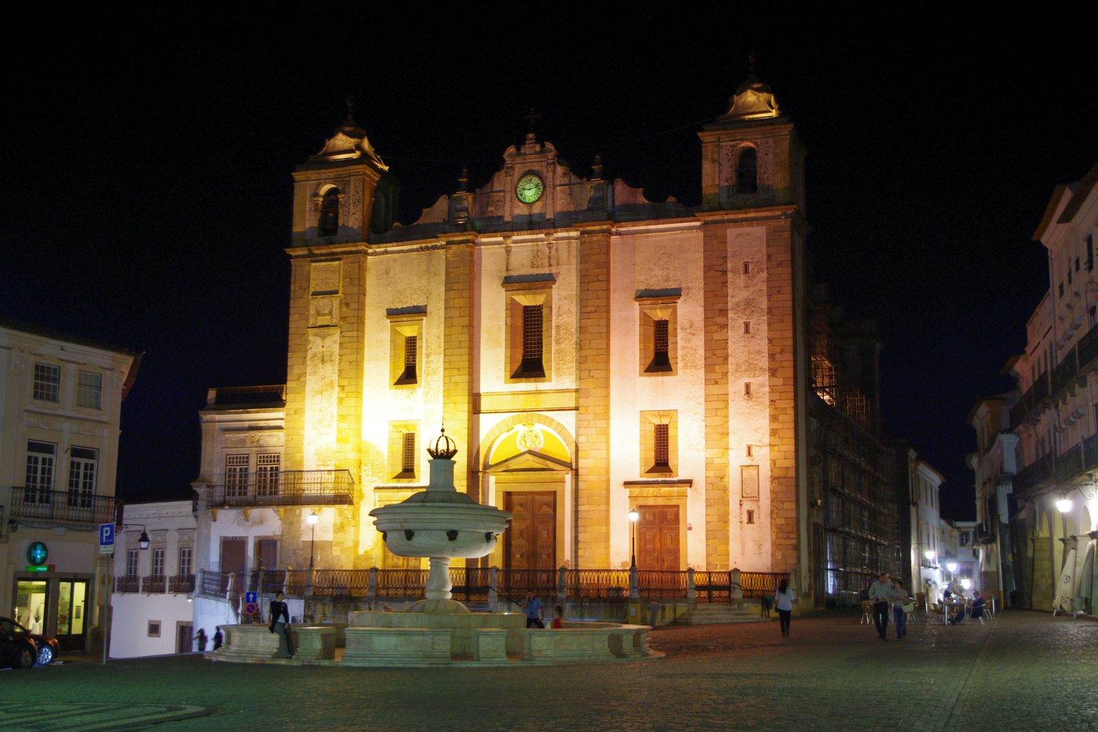 The floodlit twin towered front of the Church of Santo Antão at night, with the white marble fountain of the Praça do Giraldo before it and a few people crossing the cobbled square