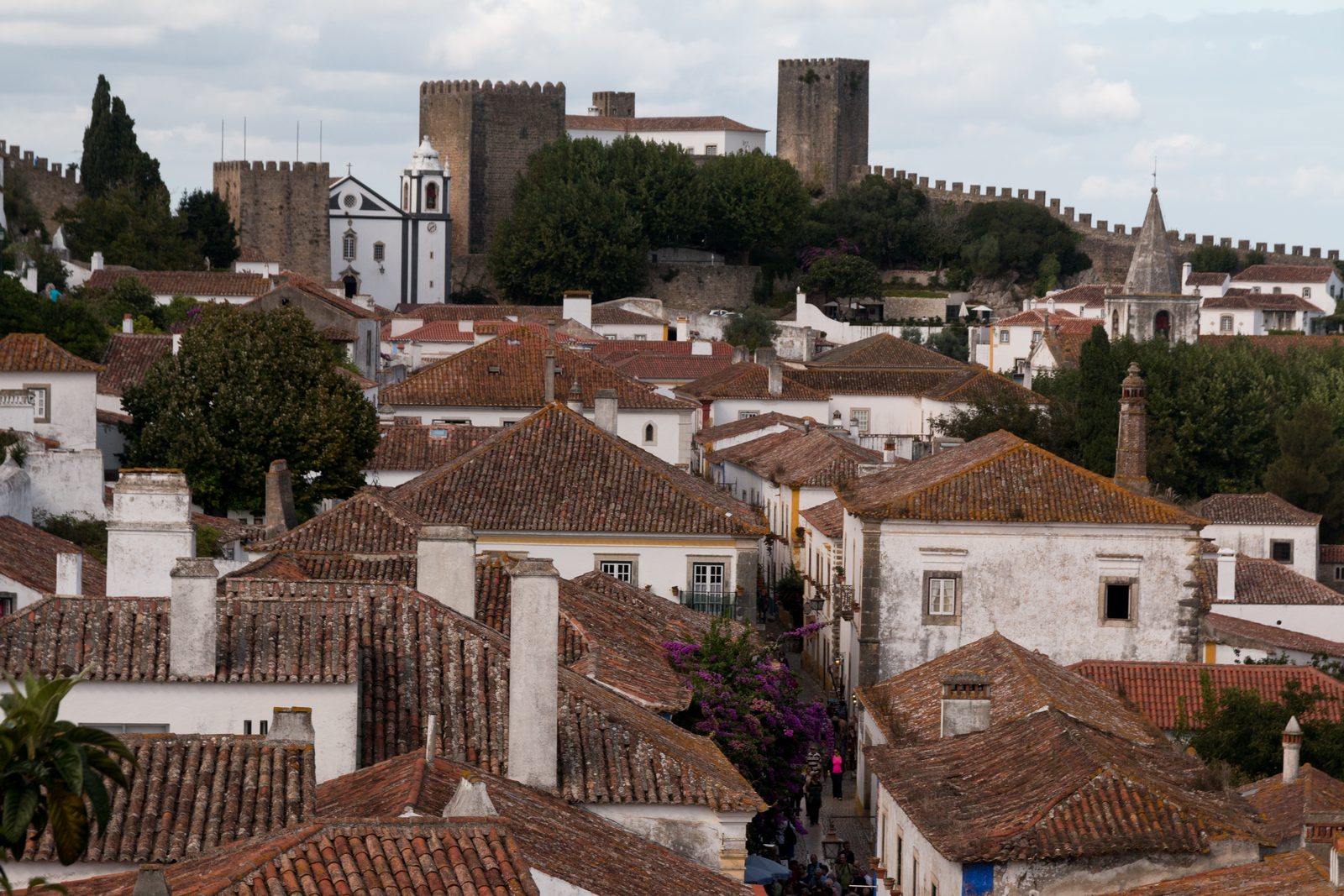 Red tiled roofs and white houses of Óbidos below the castle towers and the battlemented town walls, with a white church and its bell tower, and people walking up a narrow street lined with purple bougainvillea