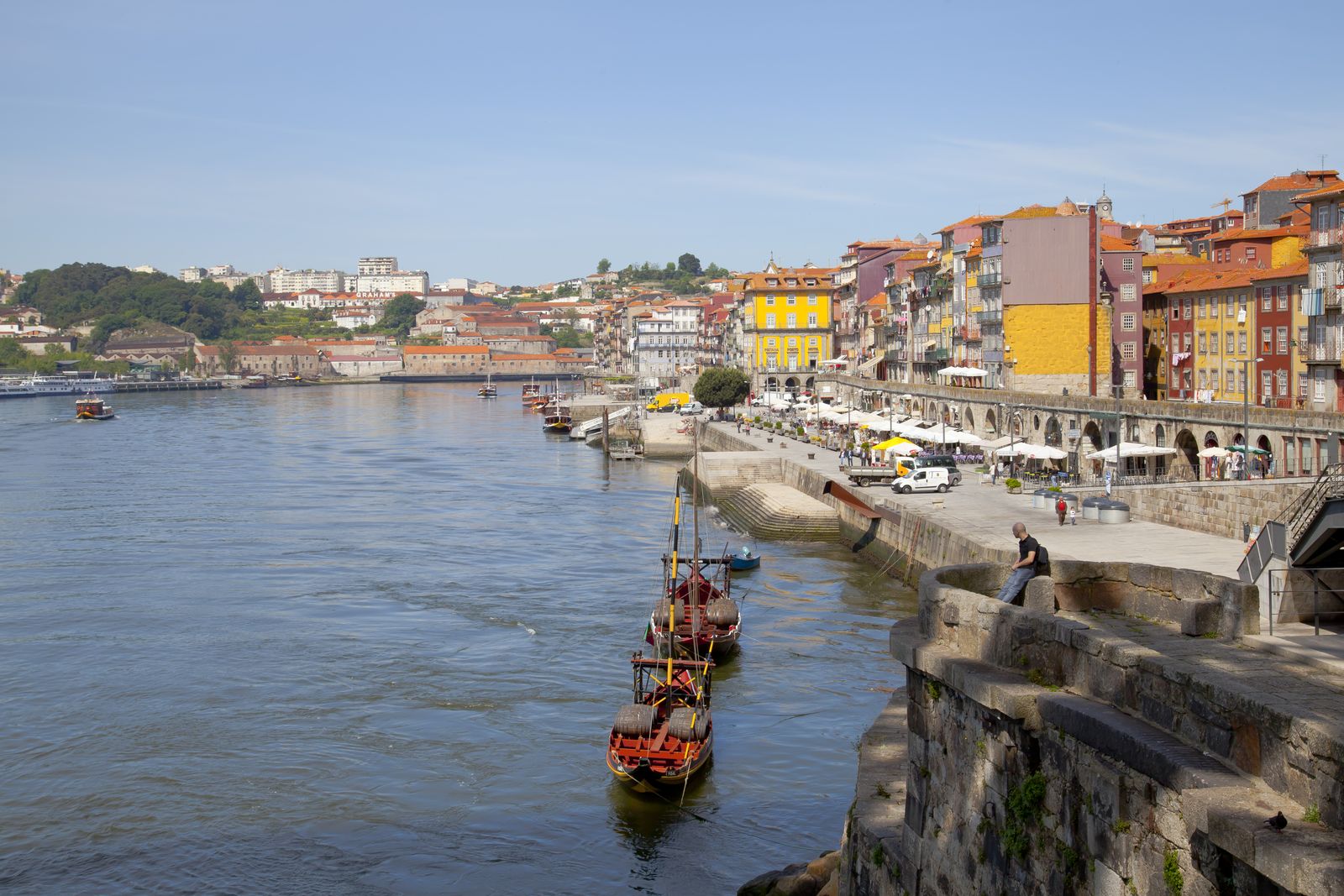 Two rabelo boats with casks on deck moored on the Douro below the Cais da Ribeira, with the arcaded quay and the tall painted houses of the Ribeira beyond