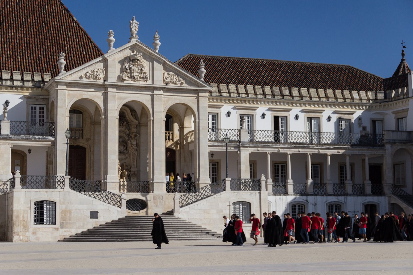 The pedimented portico and double stairway of the old royal palace at the University of Coimbra, with students in black capes and a group in red shirts crossing the courtyard below