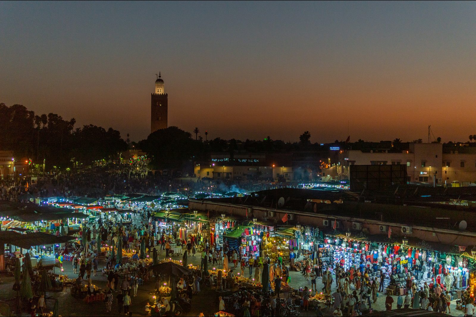 The Jemaa el Fna square at dusk, crowded with people among lit stalls and smoking food stands, with the lit minaret of the Koutoubia rising above the palm trees against an orange sky