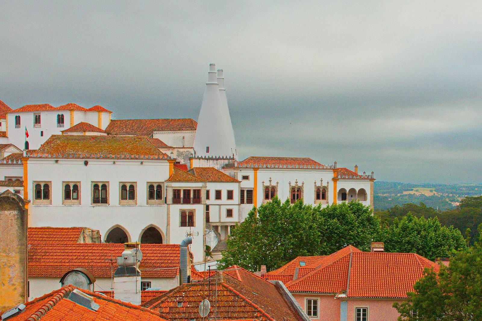 The white Sintra National Palace with its two tall cone chimneys, seen across the red tiled roofs of the old town under a grey sky, with green countryside beyond