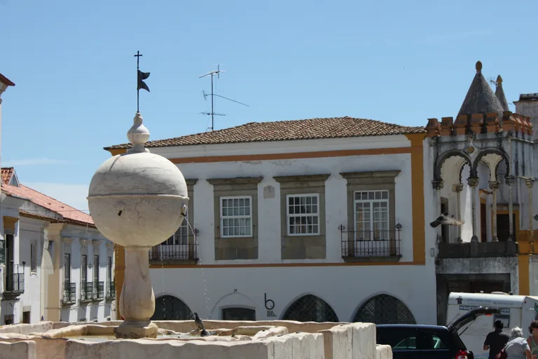 The white marble fountain of the Largo das Portas de Moura, a globe on a slender shaft, with the arched balcony of the Casa Cordovil on the right.
