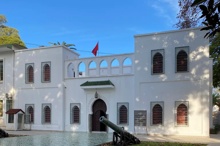 The white two storey Mendoubia with arched windows behind green grilles, an arcaded upper wall and a red flag, with an old bronze cannon on its carriage on the lawn in front.