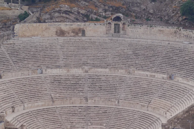 The Roman Theatre in Amman, its steep curved banks of stone seats divided by walkways and climbing to a wall with a small arched shrine at the top, cut into the rocky hillside.