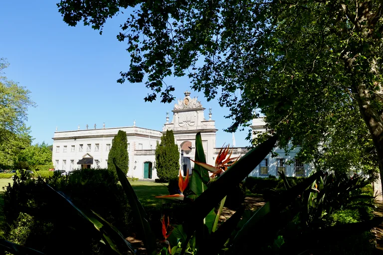 Seteais Palace seen through trees and bird of paradise flowers, with two pale wings joined by a triumphal arch crowned by a medallion.