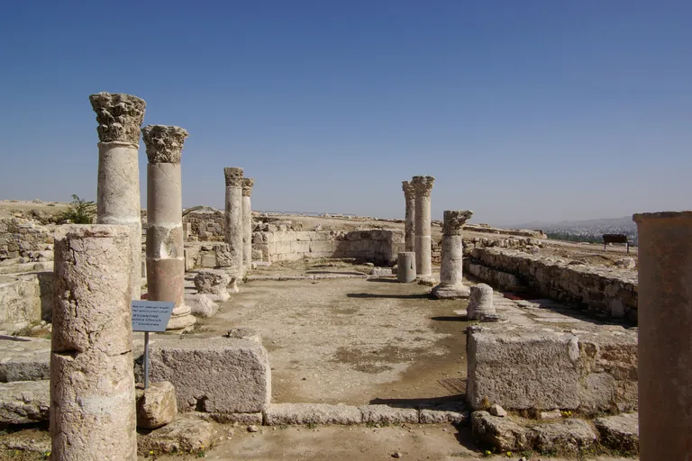 The ruined Byzantine church on the Citadel, columns with carved Corinthian capitals along the outline of the nave, flagstones and low stone walls, an information sign at the left and the hills of Amman in the distance.