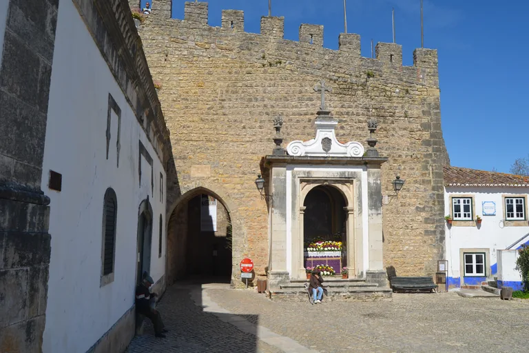 The crenellated tower of the Porta da Vila, with the arched gate passage on the left and a white baroque oratory with flowers set into the wall.