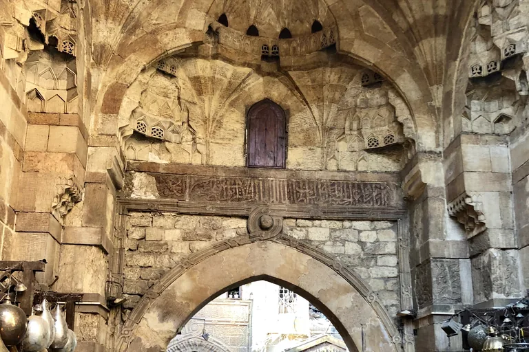 A Mamluk stone gateway in Khan el Khalili, with a vault of carved stone stalactites, a small wooden door, a band of Arabic inscription and a pointed arch below, brass lamps hanging at the edges.