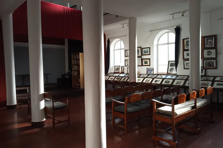 The exhibition hall of the Fondation Lorin, the former synagogue's prayer hall, with white columns, wooden benches, a raised end hung with red curtains and rows of framed photographs along the walls.
