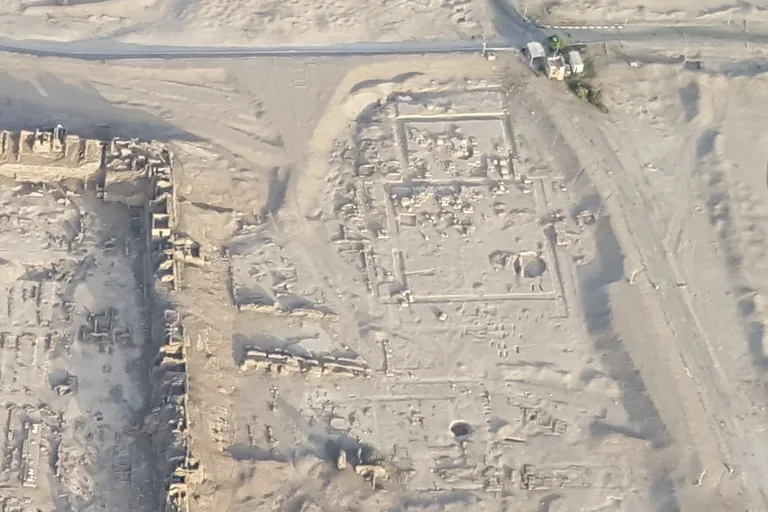 An aerial view of the low ruin field of the temple of Ay and Horemheb beside a road, the outlines of its courts and walls traced in the sand.