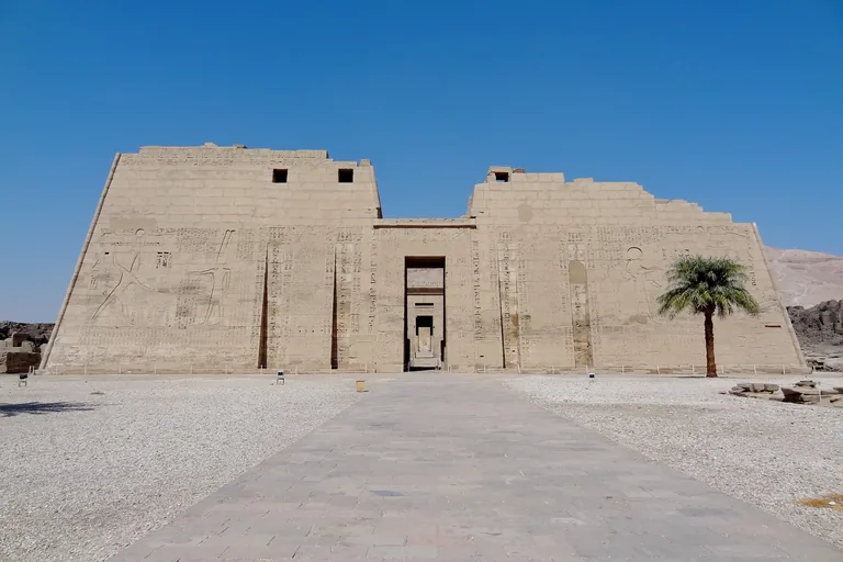 The great pylon of Ramesses III's temple at Medinet Habu, two carved towers with the king's smiting scenes and a central doorway, seen down a paved way across gravel, with a palm tree on the right.