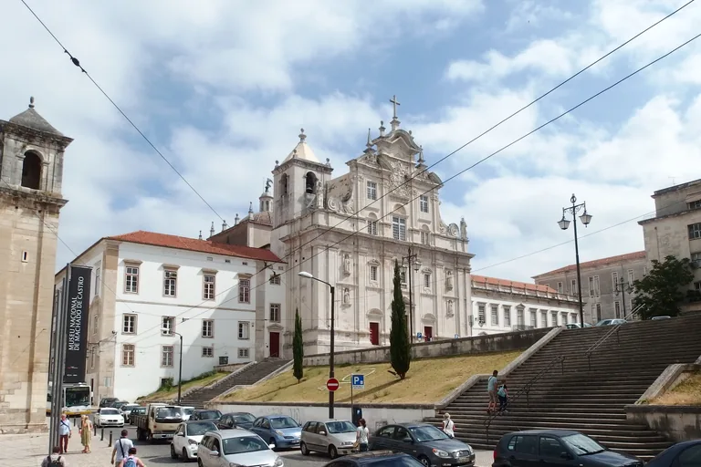 The white front of the New Cathedral with its towers and niche statues above a sloping lawn, stairs and parked cars.