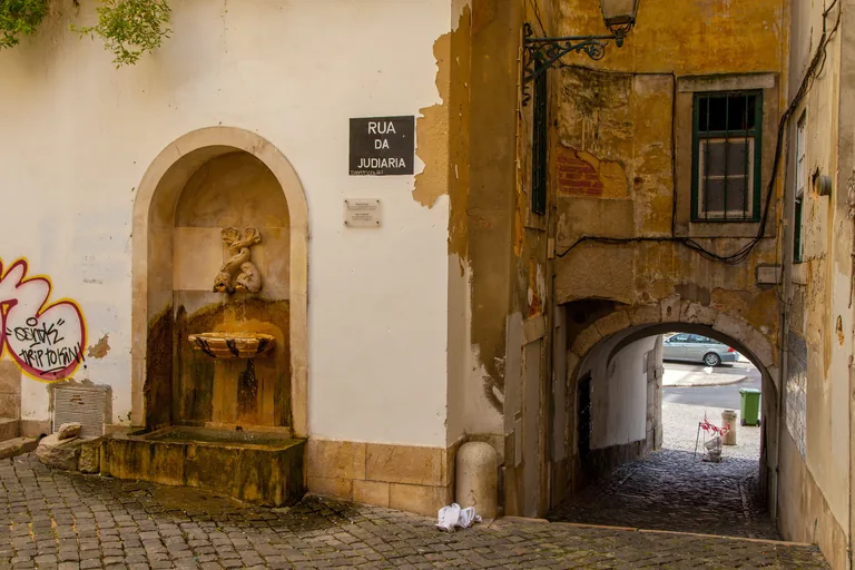 A wall fountain and an archway beneath the Rua da Judiaria street sign.