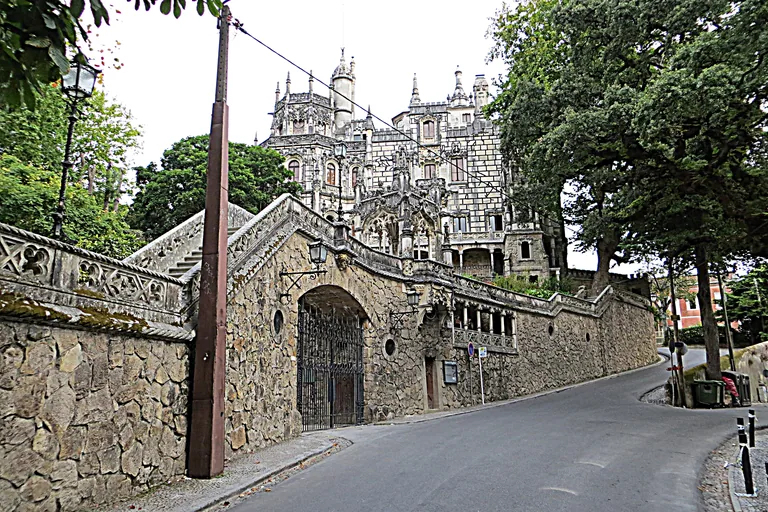 The carved towers of the Quinta da Regaleira palace rising above the estate's stone wall and iron gate on a curving road.