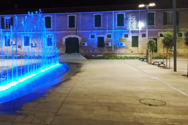 The long stone front of the former police station in Paphos at night, lit in blue, with shuttered windows and a round arched door, behind a paved square where a fountain throws jets of water lit in blue.