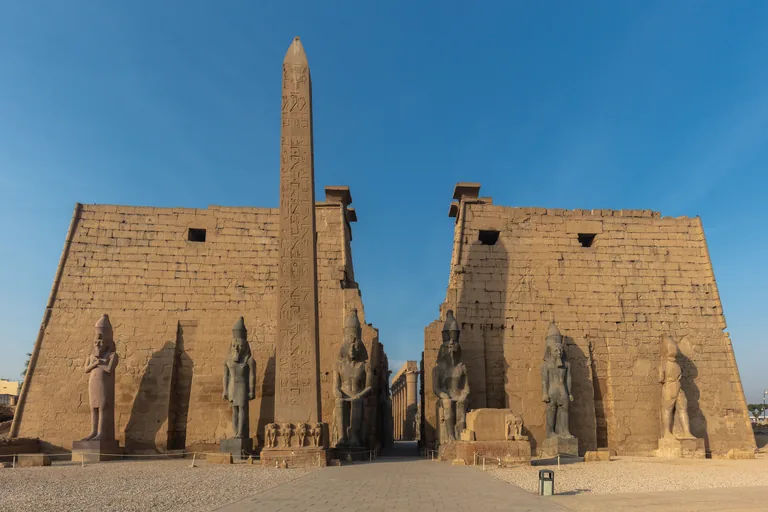 The gateway of Luxor Temple in morning light, two great pylon towers with seated and standing colossi of Ramesses II before them and a single tall obelisk standing in front of the left tower.