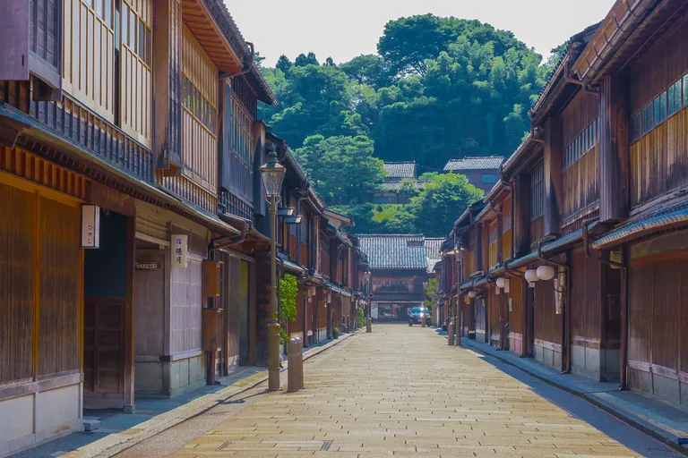 A straight stone paved street in the Higashi teahouse district of Kanazawa, lined on both sides with two storey wooden houses with fine lattices and tall upper floors, a lamp post at the left and a wooded hill rising beyond the far end.