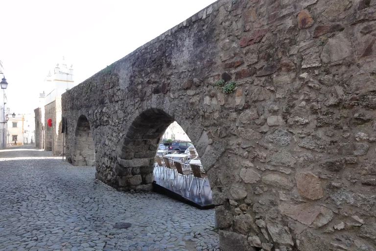The rubble stone arches of the Água de Prata Aqueduct running along the cobbled Rua do Cano, with café tables seen through one arch.
