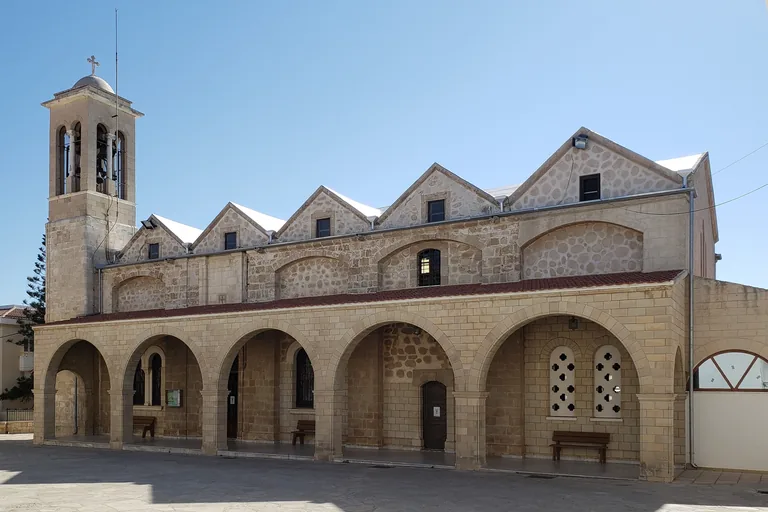 The long side of the cathedral of Agios Theodoros in Paphos, an arcade of round arches in pale stone with benches in its shade, a row of gables along the roof above and a square bell tower at the left end.