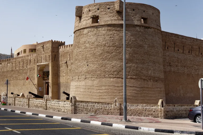 The east side of Al Fahidi Fort, a broad round tower and crenellated walls of plastered stone above a low stone wall along the pavement, with cannons and flags at the gate.