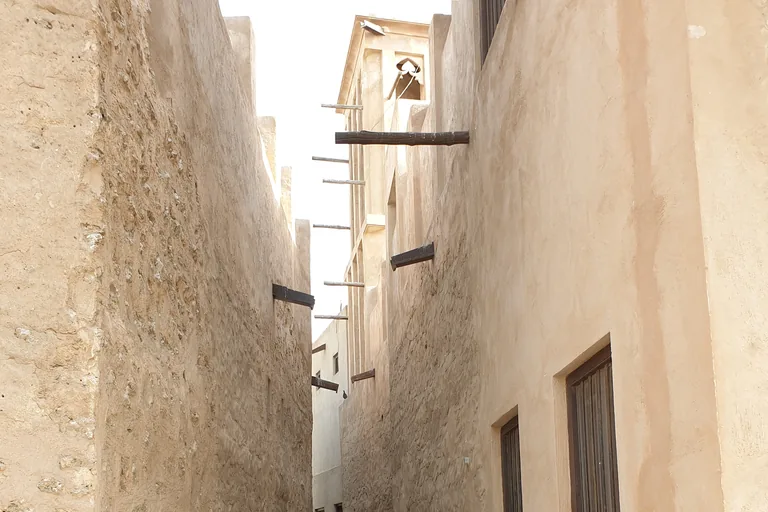 A narrow lane between high walls of rough stone and smooth plaster, with wooden beams jutting from the walls and a square wind tower above the far end.