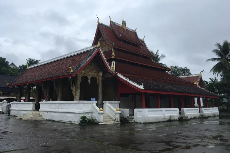 The hall of Wat Mai in Luang Prabang, a long building with roofs of dark red tiles falling in tiers, gilded finials and a porch of carved gilded pillars, on a white plinth with steps, seen across a wet paved court under a grey sky.
