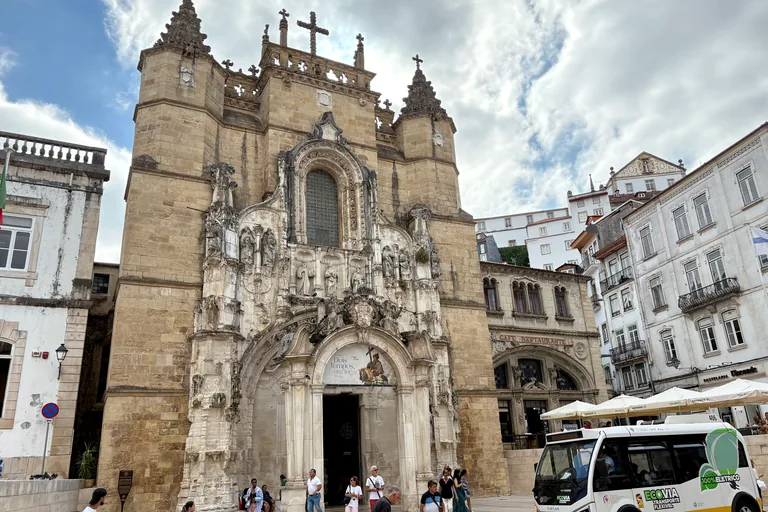 The Manueline front of the church of the Monastery of Santa Cruz, with twin turrets and a carved portal on a paved square, and a café's arched windows to the right.