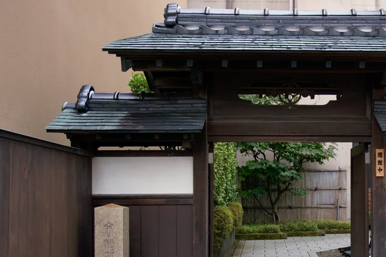 A small dark wooden gate with tiled roofs, a white plaster panel and a stone post carved with characters at the left, opening onto a paved path and a garden with a maple and a bamboo fence, at the Izumi Kyoka Memorial Museum in Kanazawa.