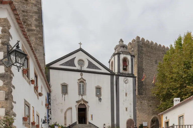 The white front of the church that houses the Livraria de Santiago, with its bell tower and steps up to the door, and a crenellated castle tower behind.