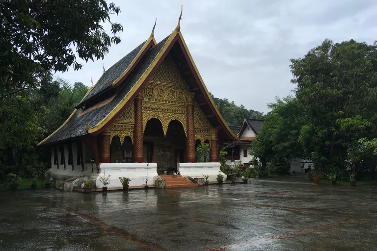 The hall of Wat Xieng Muan in Luang Prabang, a steep roof of dark tiles over a gable and porch covered in gilded carving with red and gold pillars, on a white base with steps, behind a wet paved court and trees.