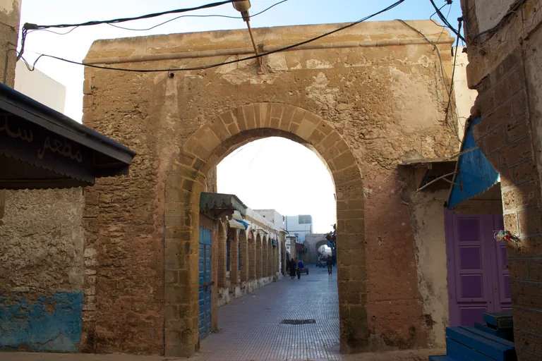 A round stone arch over a paved market street leading into Souk Jdid, with shuttered shops under arcades along the street beyond.