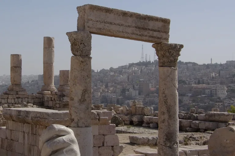 Two columns of the Great Temple on the Citadel carrying a carved stone beam, with column stumps and fallen blocks on the platform, part of a colossal marble hand at the lower left and the houses of Amman on the hills beyond.