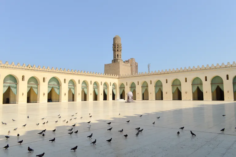 The white marble courtyard of the mosque of al Hakim under a clear sky, ringed by arcades hung with pale green curtains and crowned with stepped crenellations, with a minaret rising from a square stone bastion and pigeons on the paving.