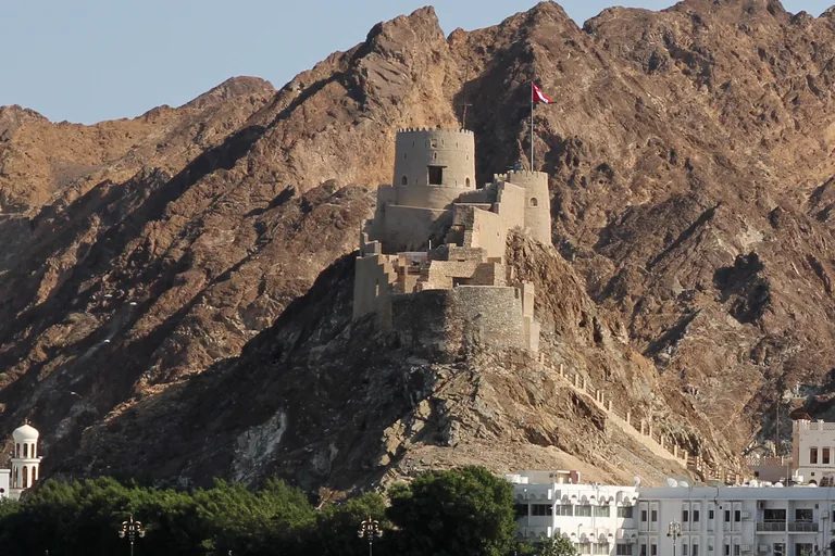 Mutrah Fort on its rocky ridge above the town, round stone towers and walls stepping down the brown rock, with white buildings and trees at the foot of the hill.