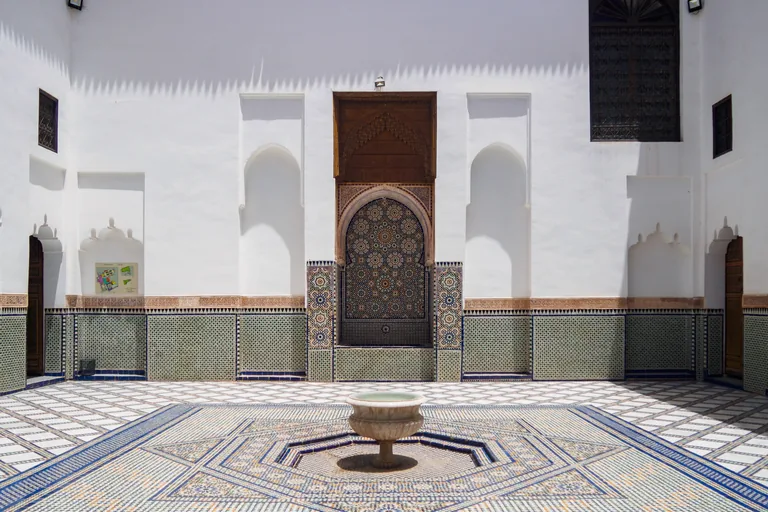 The white walled courtyard of Dar Si Said with a marble basin on a geometric tiled floor, green and blue tiled dados and a tiled fountain niche under a carved wooden canopy.