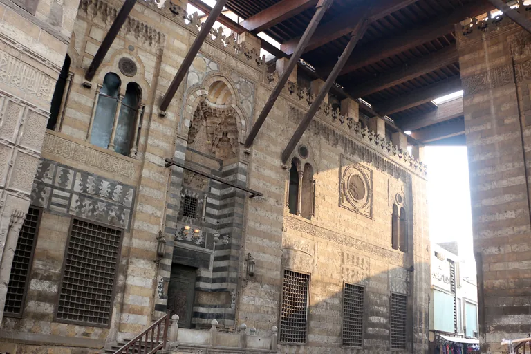 The striped stone front of the al Ghuri mausoleum under the timber roof that spans the street between the two halves of the complex, with tall grilled windows and a carved doorway.