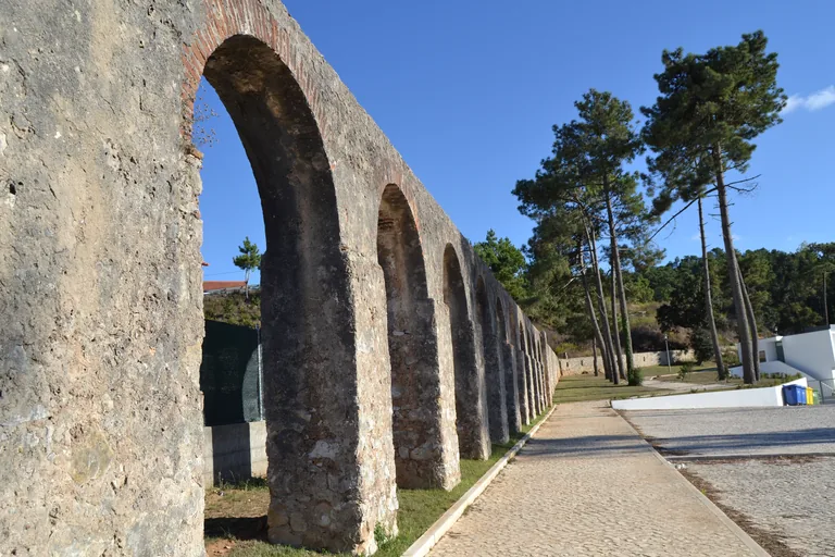 The stone arches of the Óbidos Aqueduct running beside a paved path and a lawn, with pine trees and a white house to the right.