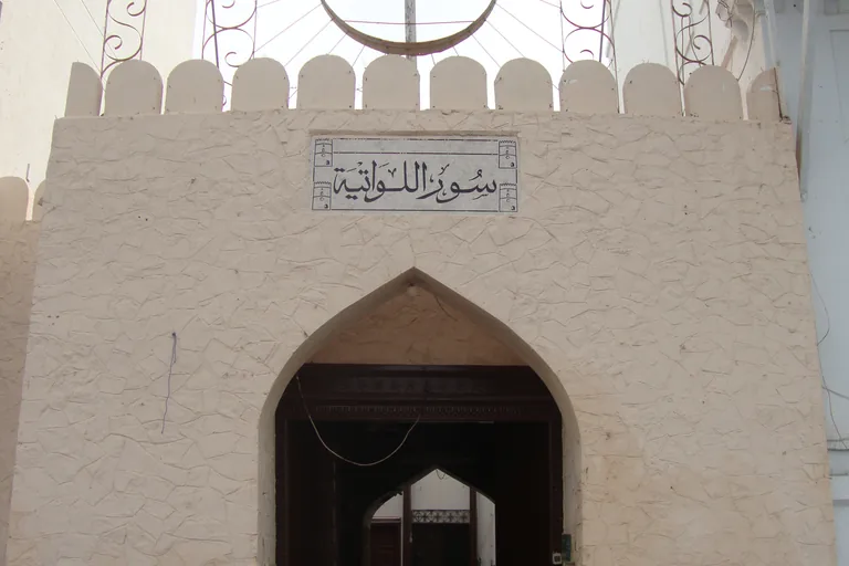 A gate of Sur al-Lawatiya, a pointed arch in a whitewashed crenellated wall under an Arabic name plaque, opening onto a dark passage and an inner arch.