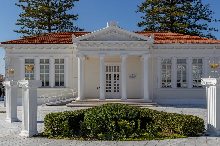 The Municipal Library of Paphos, a white building with a pediment on four Ionic columns under red tiled roofs and two tall pines, with white columns carrying golden bowls and a round hedge in the paved square in front.