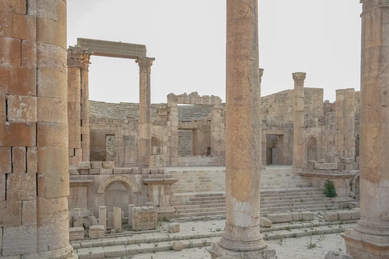 The North Theatre at Jerash seen between tall columns of the street, its stage front with columns and doorways, broad stone steps and the curved rows of seats behind.
