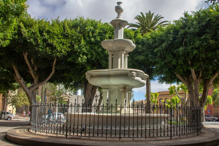 A white marble fountain with tiers of basins held on thin columns inside a round iron railing in the Plaza del Adelantado, under the spreading trees of the square, with painted house fronts and a palm beyond.