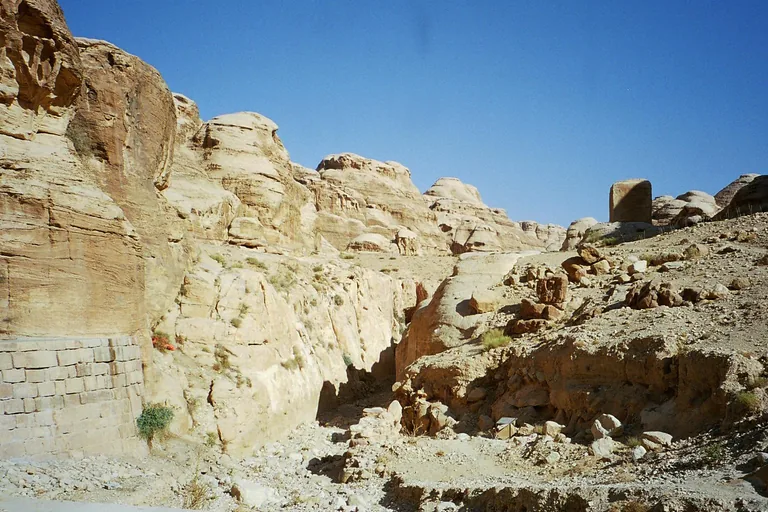 The approach to the Siq, a stone retaining wall on the left and a dry rocky channel leading into a cleft between pale sandstone cliffs, with a block monument standing on the slope at the right.