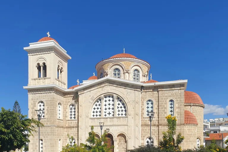 The church of Agios Kendeas in Paphos, pale stone with a square bell tower, a central dome and smaller domes roofed in red tiles, under a deep blue sky with trees in front.