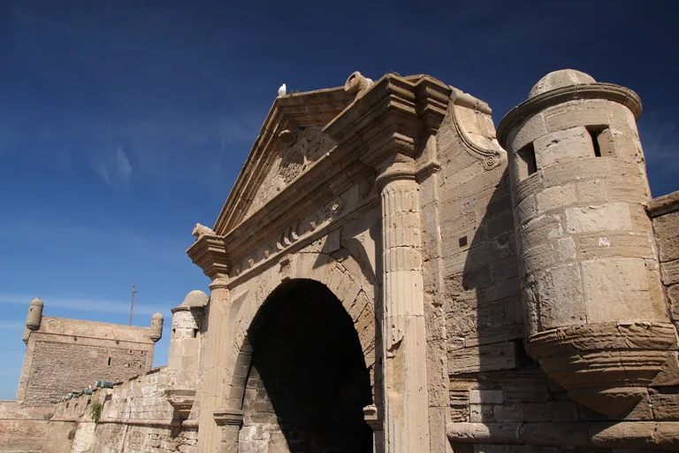 The carved stone face of Bab el Marsa, with a triangular pediment, fluted columns and a round corner turret, under a deep blue sky with the port bastion behind.