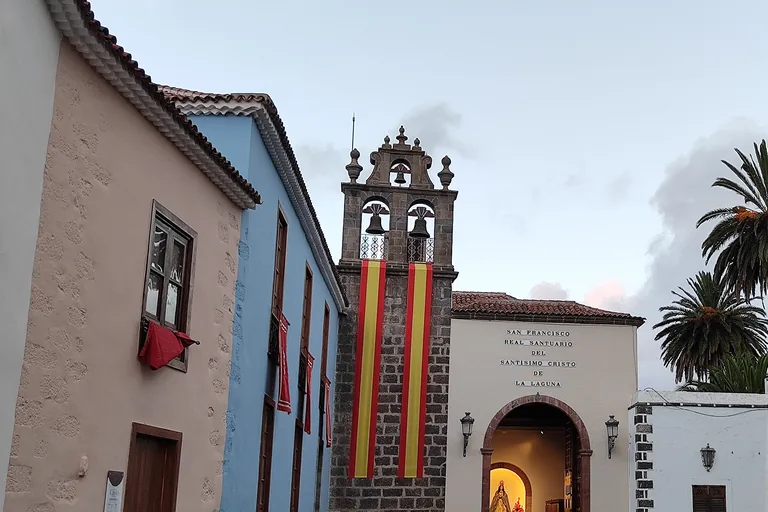 The sanctuary of the Cristo seen past a pink and a blue house front, a dark stone bell gable with three bells and long red and yellow banners beside a white front lettered with the sanctuary's name over an arched doorway.