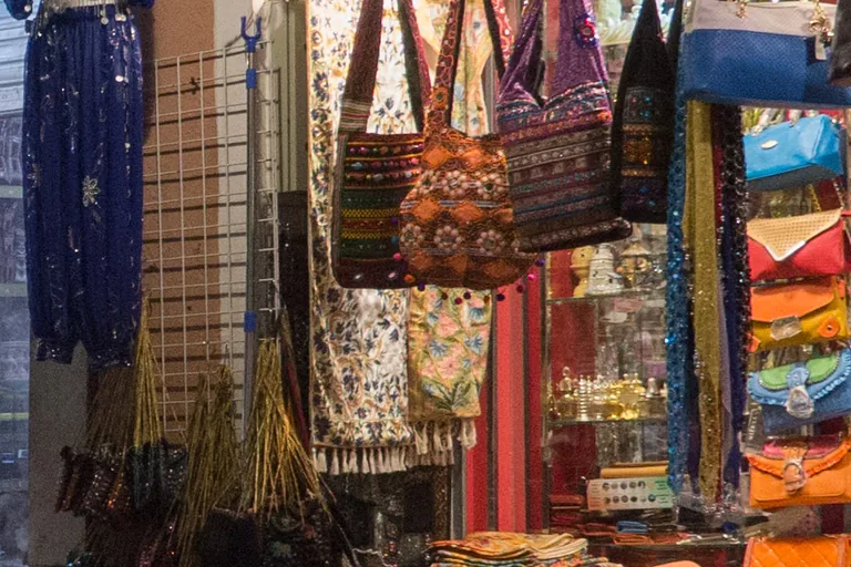 Embroidered cloth bags, scarves and a dark blue costume hanging at the front of a shop in a covered lane of the Mutrah souq, with handbags and folded cloth below.