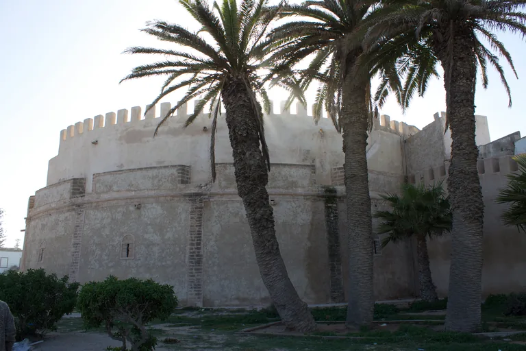 The round crenellated bastion beside Bab Marrakech, its pale walls behind tall palm trees and a lawn.