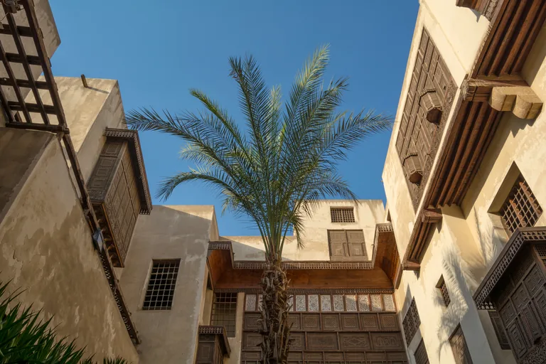 The courtyard of Bayt al Suhaymi seen from below, a date palm rising between pale plastered walls with projecting turned wood window screens and a roofed wooden loggia under a blue sky.