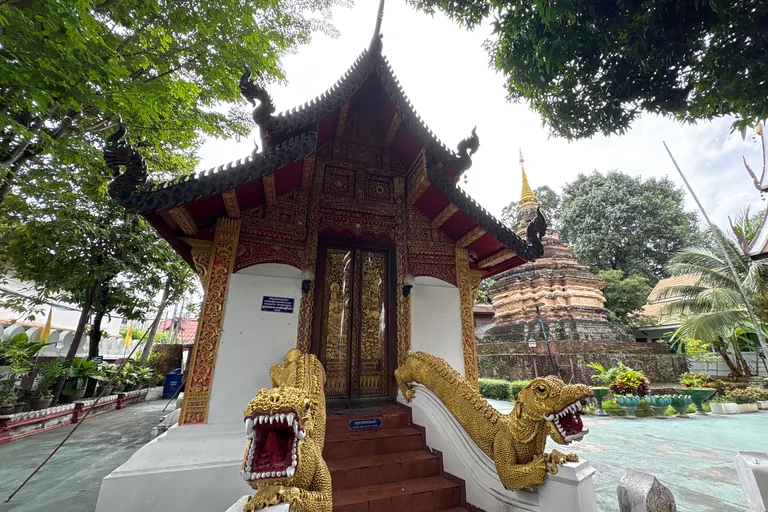 The small hall of Wat Umong in Chiang Mai, its steep dark roof with curling finials over a red and gilded carved front and gilded doors, two golden guardian beasts with open jaws on its stair, and an old brick chedi with a gilded spire among trees behind it.